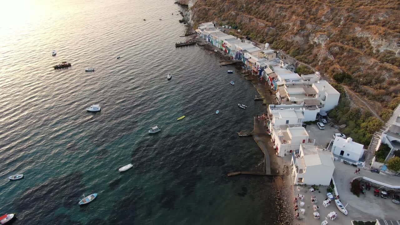 vista de dron en grecia volando sobre una pequeña ciudad con casas blancas y puertas de colores al lado del mar y la montaña en milos al atardecer