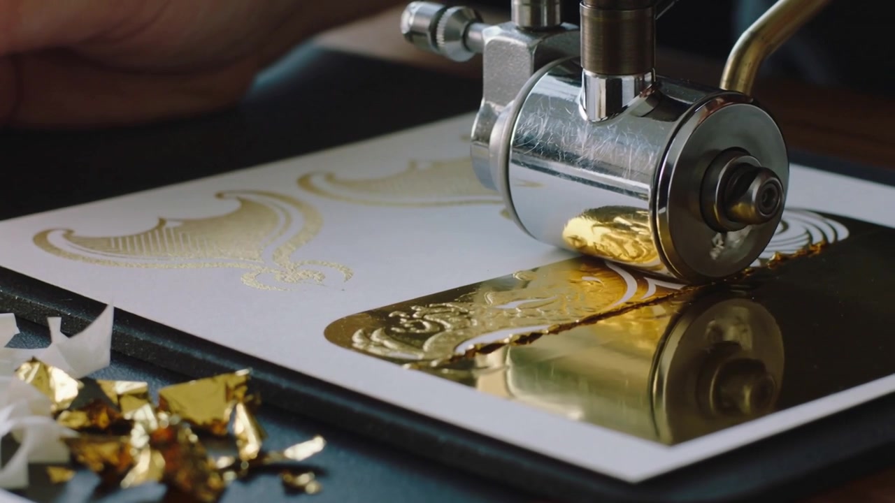 Rolling artisan hand dark-sleeve pressing foil roller across cardstock revealing ornate gold design