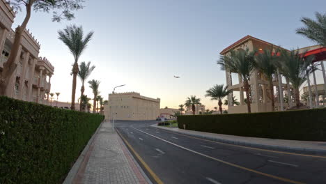 An-avenue-between-monumental-houses-in-a-tropical-location-with-palm-trees,-a-plane-passes-in-the-blue-sky