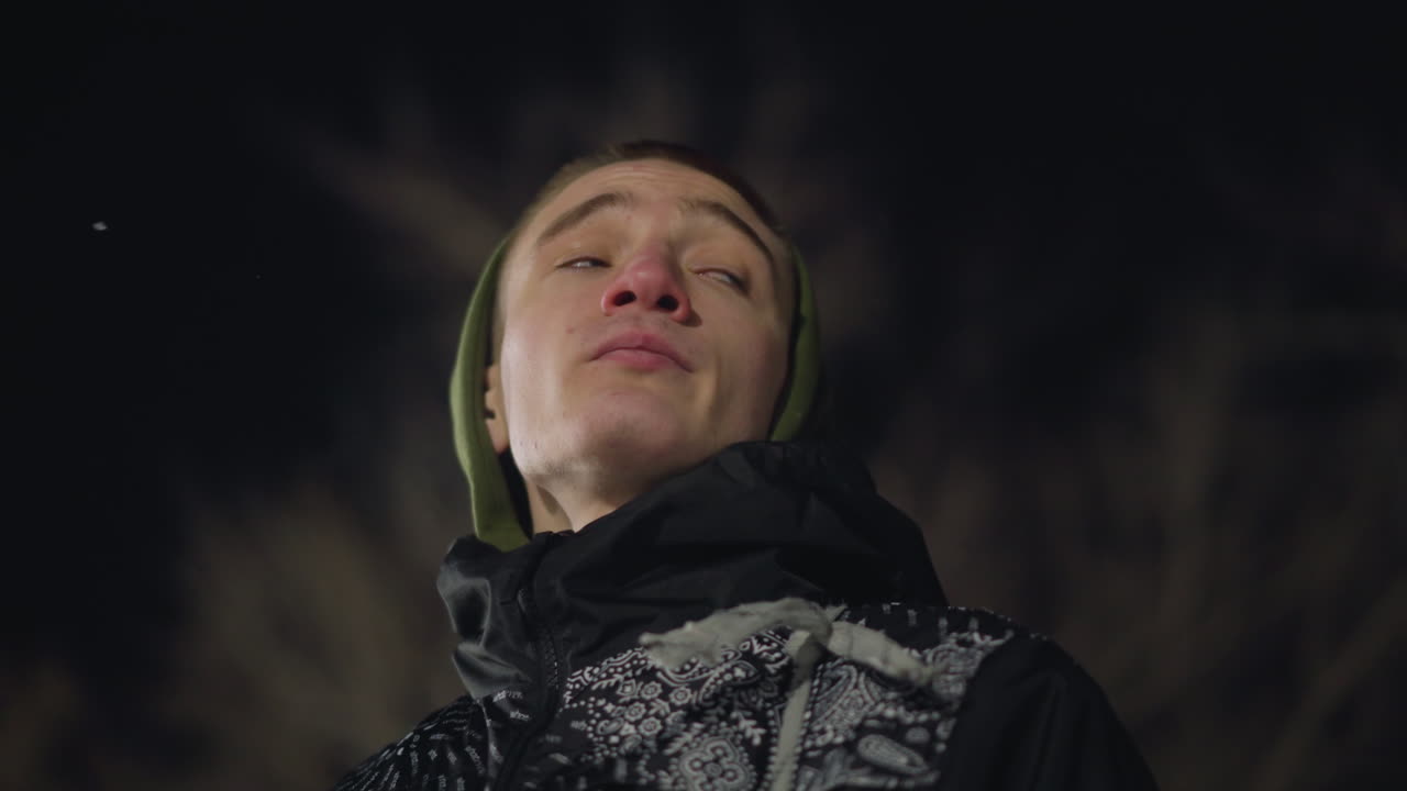 Close up of man in green hoodie and black jacket looking around contemplatively during cold winter night, breath forming mist, bare tree branches blurred in background under soft light
