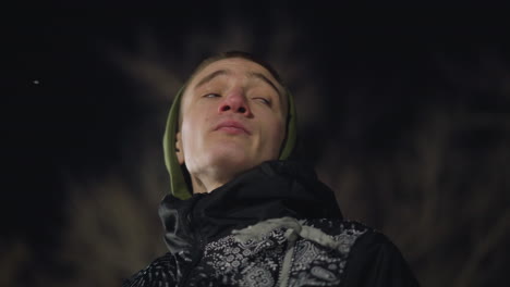 close up of man in green hoodie and black jacket looking around contemplatively during cold winter night, breath forming mist, bare tree branches blurred in background under soft light