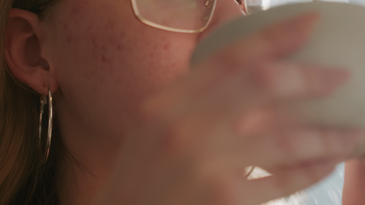 Donna bianca che sorseggia caffè alla luce del sole, primo piano di donna caucasica con lentiggini che indossa occhiali e orecchino a cerchio, sorriso gentile, tazza di ceramica, bagliore cinematografico caldo, rituale accogliente in cucina, bokeh morbido