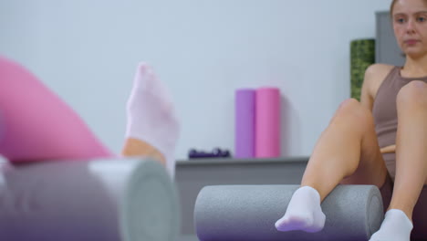 dos mujeres practicando ejercicios para las piernas con rodillos de espuma en esteras de yoga en un gimnasio bien iluminado, centrándose en la flexibilidad y la recuperación muscular, con equipos de gimnasio sutilmente visibles en el fondo