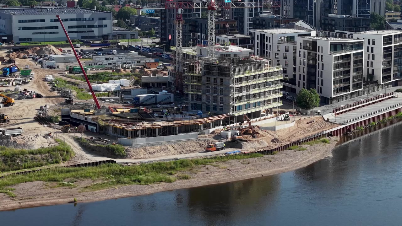 Luchtopnamen tonen actieve bouw langs de rivier met kranen, moderne appartementencomplexen, druk materieel en helder weer, wat stedelijke groei en ontwikkeling benadrukt