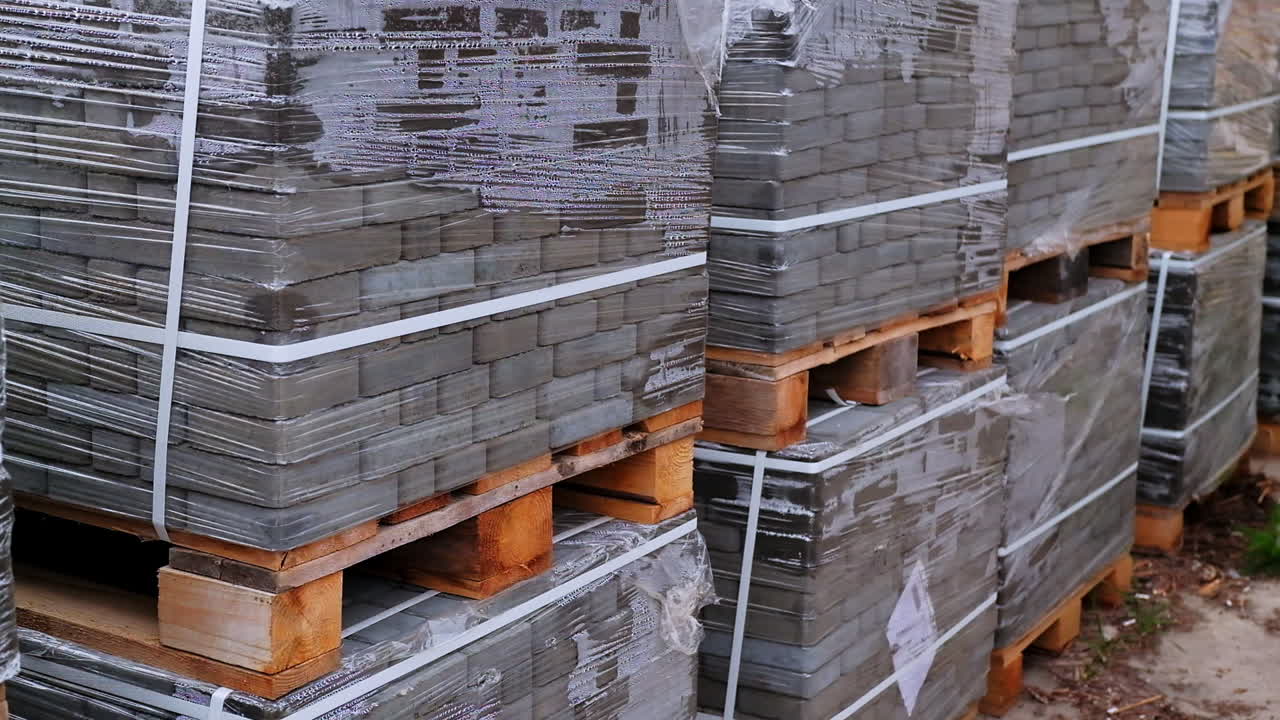 Huge packs of grey bricks stacked at wooden pallets. Construction materials covered with cellophane and tied with plastic band.