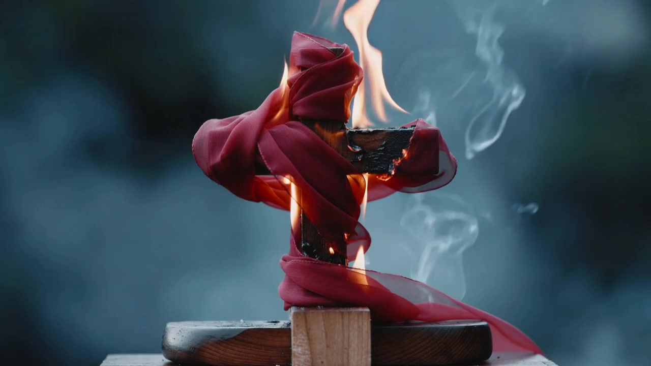Una fiamma si accende, incendiando una croce di legno avvolta in un panno rosso posta su un blocco all'aperto, con il fumo che si sposta verso destra.