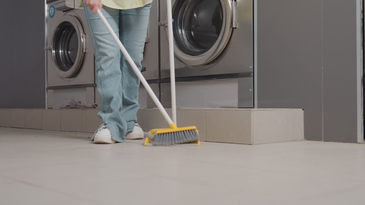 Janitor packs dirt from floor with broom stick and dust pan near industrial washers in laundromat, collecting lint from filter drawer spill into pan, routine cleanup, maintenance, hygiene, smiling