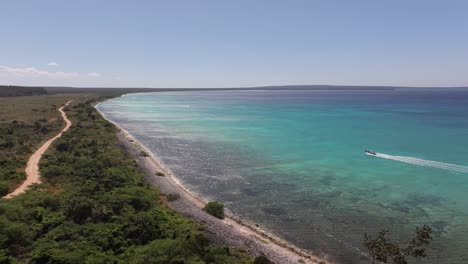 Drone-ascends-dry-tropical-vegetation-and-rocky-shore-as-boat-drives-along-in-deep-blue-water-off-shore