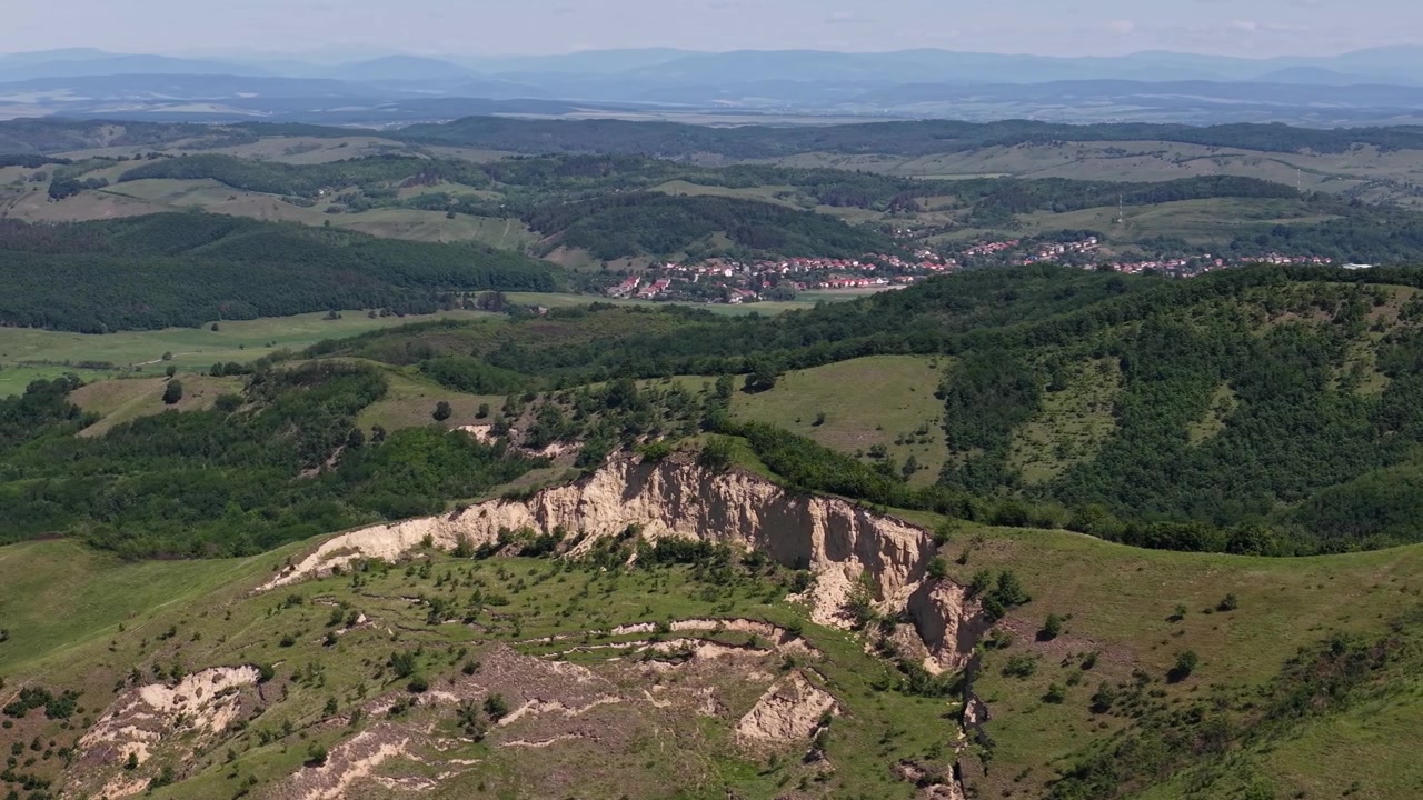 Una drammatica formazione geologica mostra l'erosione su un versante verde, con un lontano villaggio rurale annidato nella valle