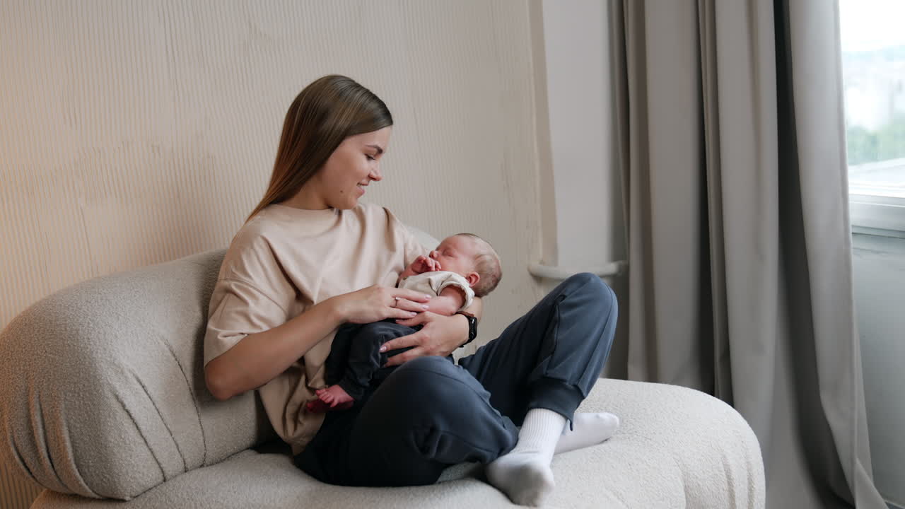 Young long-haired woman sitting on the sofa holding her tiny baby. Little infant is sleeping while mom waving him.
