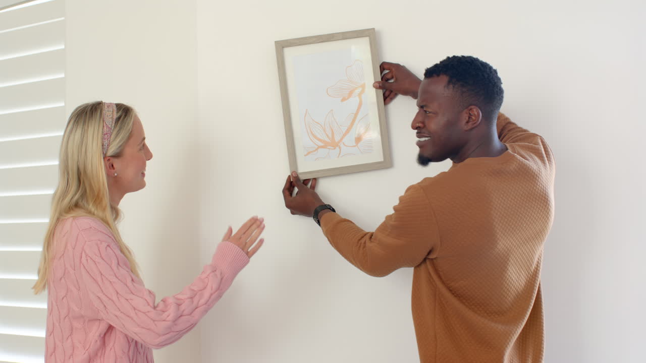 diverse couple hanging artwork on wall, smiling and enjoying home decorating together
