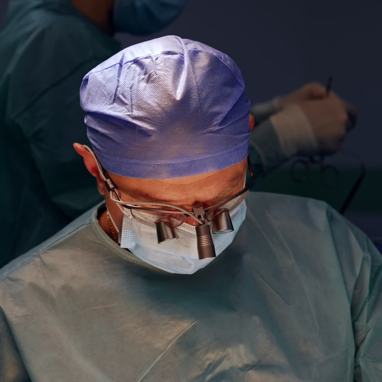 Chirurgo uomo con occhiali da chirurgo, mascherina e cuffia. Il medico è seduto accanto al paziente e osserva la zona operata.