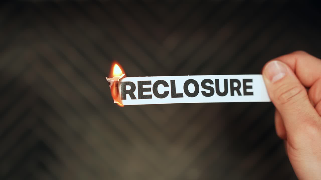 Close-up of a hand holding a paper strip labeled “FORECLOSURE” as the edge burns with flame on a dark background. Mortgage default and housing crisis concept with copy space