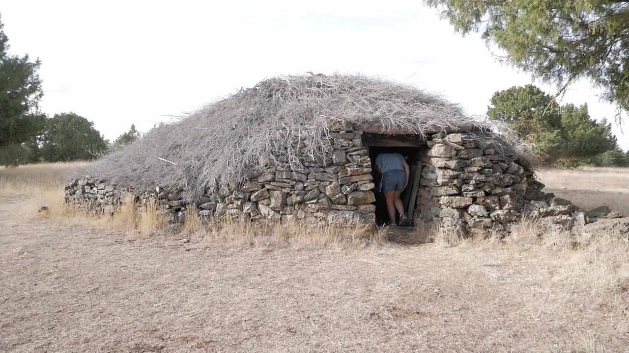 Une femme s'approche d'une vieille cabane en pierre au toit végétalisé dans un paysage rural sec, scène de jour.