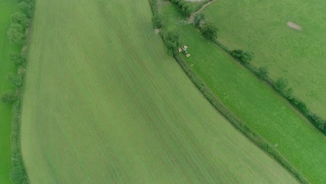 aerial tracking along green english fields with a larger tractor and mower cutting the grass in a narrow field