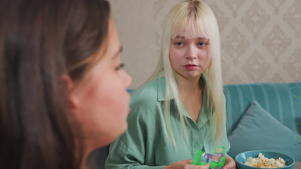 Due amici seduti a tavola che condividono snack, uno tiene una confezione verde annusandone il contenuto, l'altro con una ciotola di popcorn sorridente, in un ambiente interno accogliente con un momento sociale allegro e un'atmosfera rilassata.