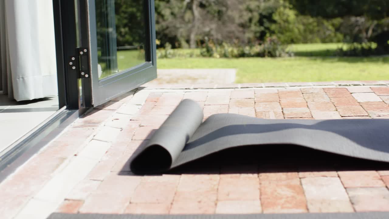 Smiling friends sitting on brick patio initiating workout by unrolling black yoga mat toward garden