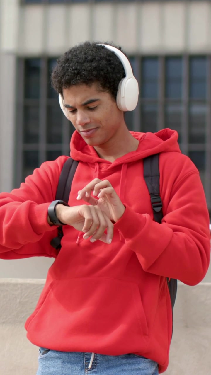 Vertical video: Adjusting headphones, red-hoodie man tapping smartwatch on rooftop, starting music
