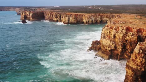 eroded sea cliffs of praia do tonel beach in portugal splashed by strong ocean waves, aerial flyover reveal shot