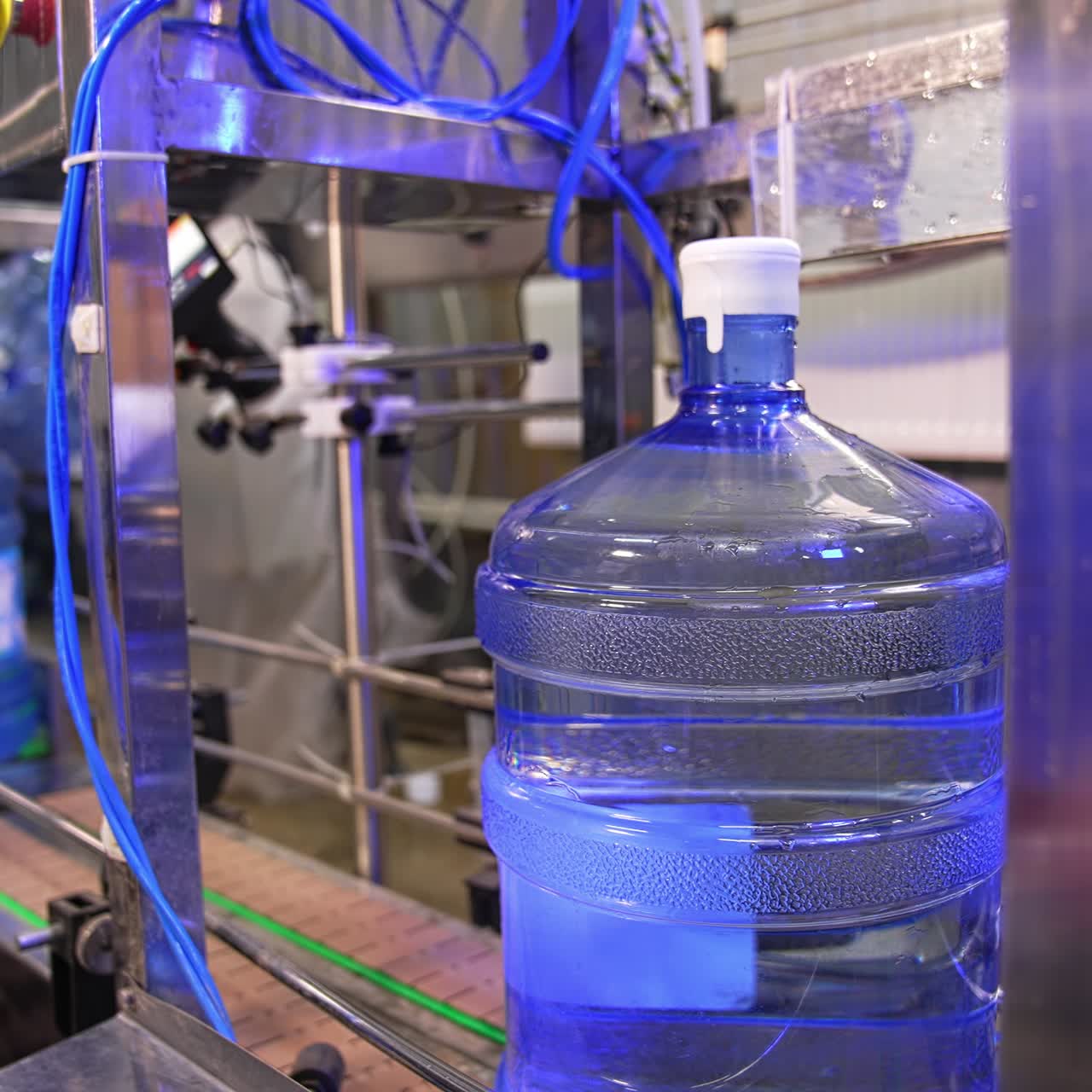 Big full bottles of clean water move by the conveyor belt. Machine is lifting a bottle and worker covers it in plastic bag