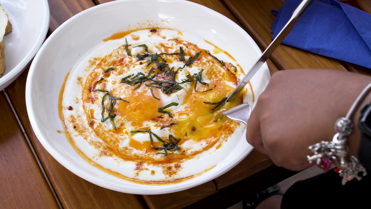 Close-Up of Hands Enjoying Cilbir Turkish Eggs in White Bowl with Flatbread at Cafe Setting