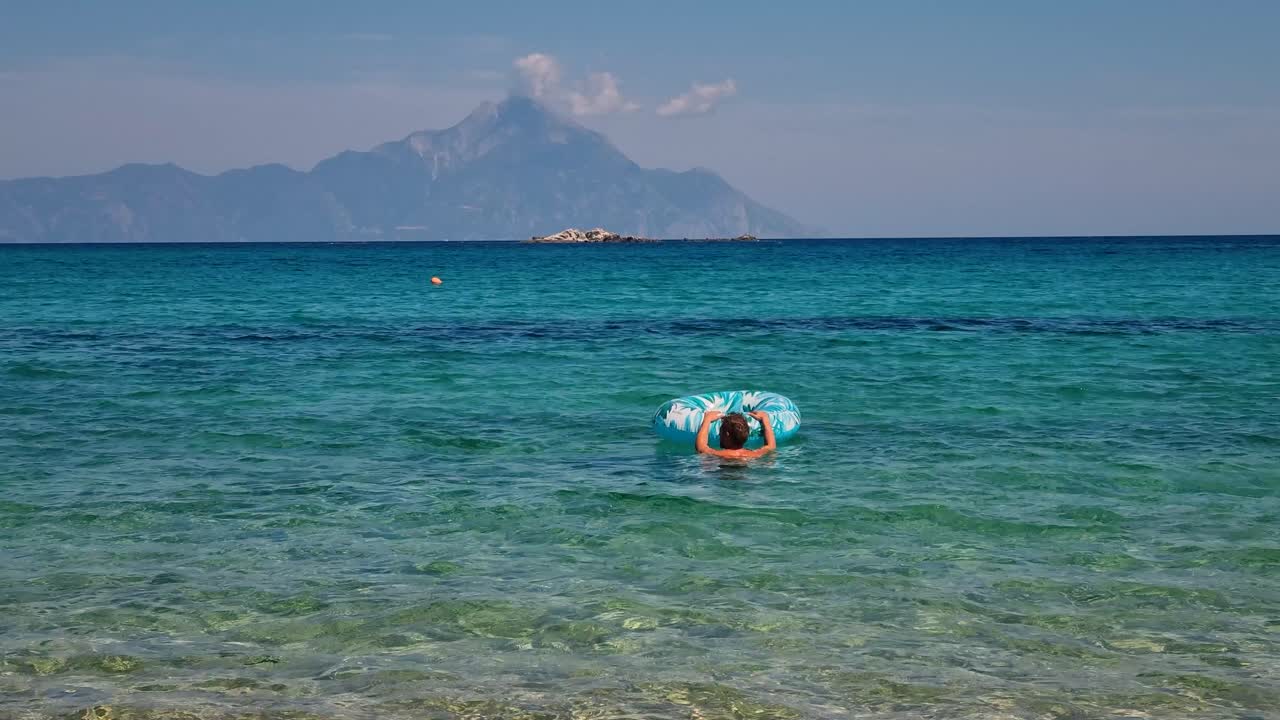 Man floating with an inflatable ring in the crystal-clear turquoise waters of Kriaritsi, Greece, enjoying a sunny Mediterranean summer holiday
