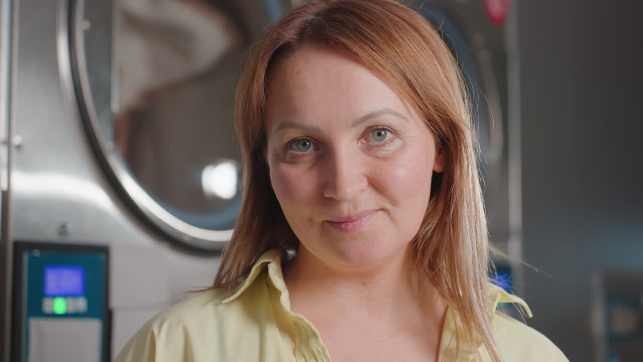 Portrait of business woman raising head up standing before industrial dryer and washer inside modern laundromat, stainless drums polished, digital panels glowing, confidence, hygiene commitment