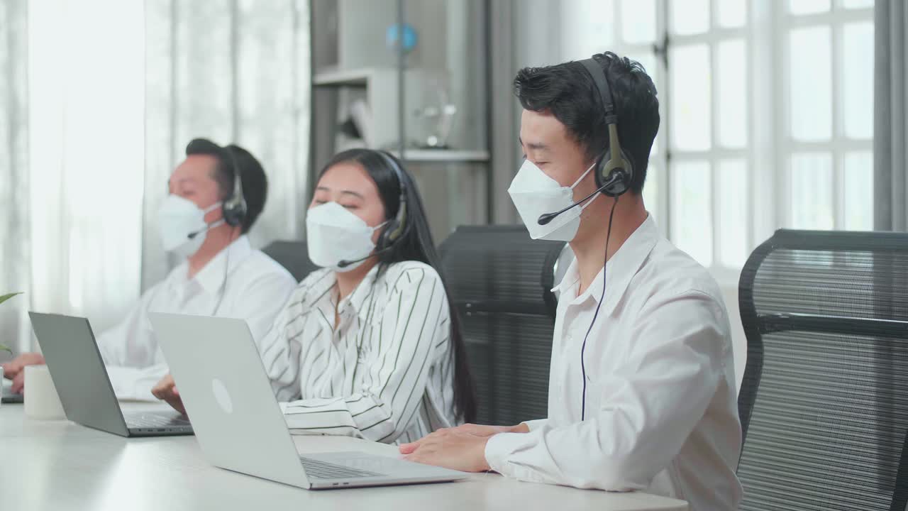 Three Asian Call Center Agents Wearing Headsets And Masks Being Happy Due To Success Working With Computers At The Office