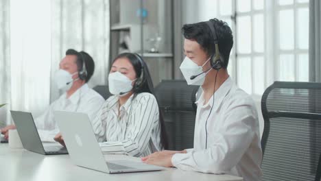 three asian call center agents wearing headsets and masks being happy due to success working with computers at the office