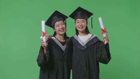 asian woman students graduate in caps and gowns hugging each other, smiling, and showing diplomas in their hands to camera on the green screen background in the studio