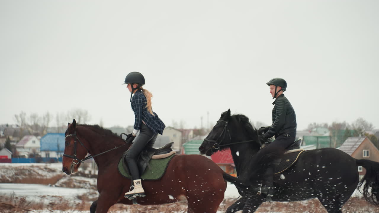 Duo caucasico che cavalca cavalli in tandem in un campo innevato imparando abilità equestri, studentessa e istruttore maschio con caschi, selle e briglie verdi, case del villaggio sullo sfondo, educazione calma.