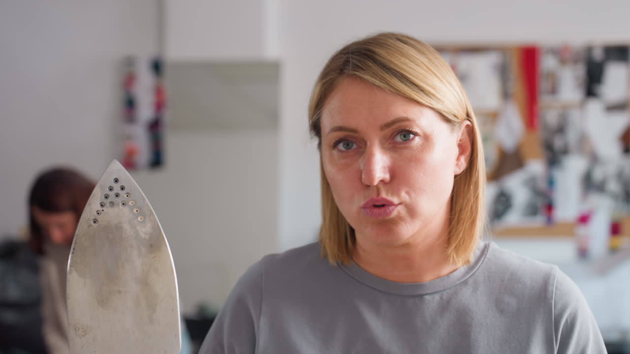 Female tailor lifts steam iron, playfully making funny face, positive energy in bright workspace, comedic vibe, focusing on skillful craft, showing professional approach with lighthearted flair