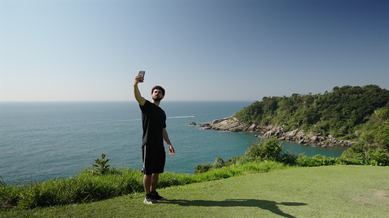 Young traveler stands on lush coastal cliff takes selfie with sparkling ocean backdrop