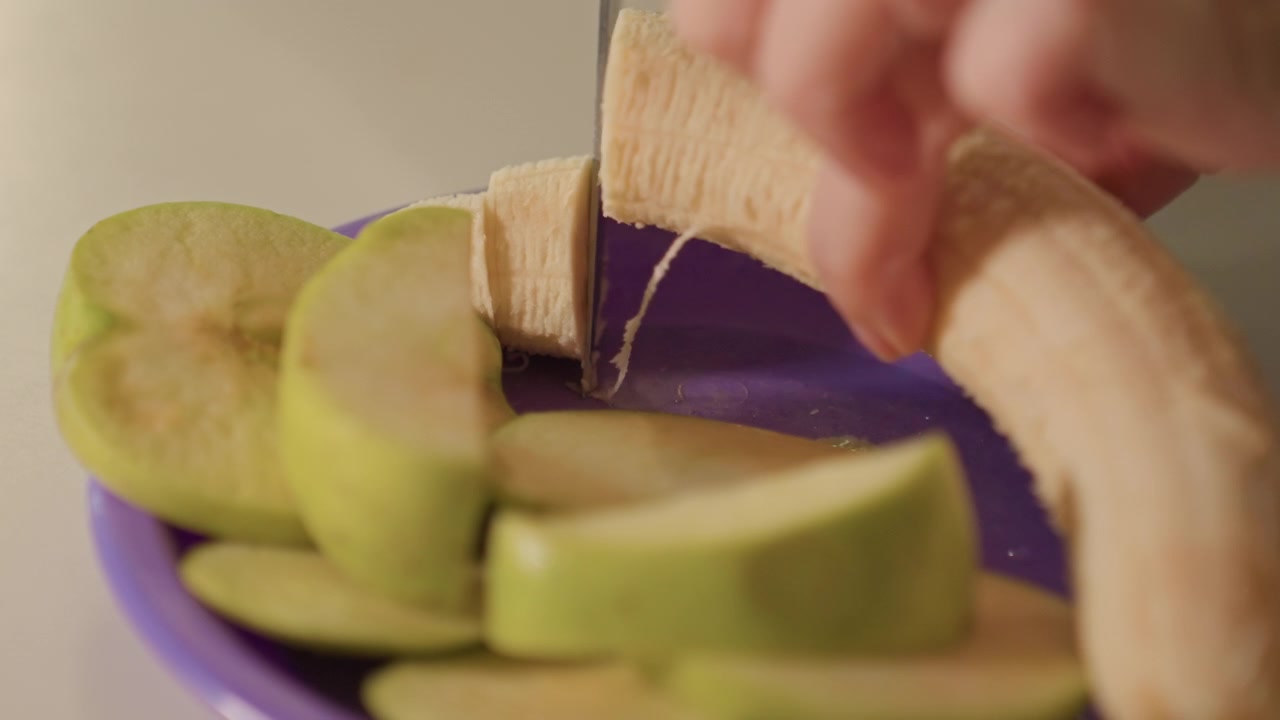 Primo piano di una mano che affetta mele verdi e banane su piatti viola. Il coltello si muove costantemente attraverso la frutta creando fette sottili. Lo sfondo semplice enfatizza la preparazione del cibo.
