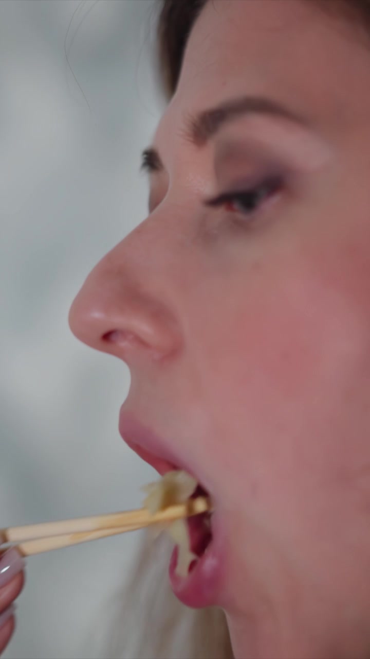 Close up of woman eating food with chopsticks focusing on her mouth and facial expressions. scene shows texture detail of skin under soft lighting as woman moves slightly while chewing