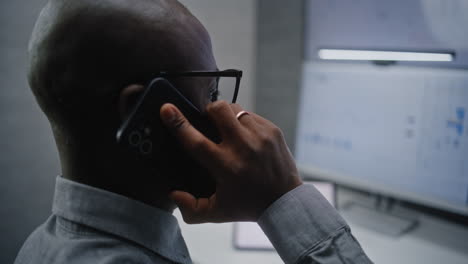 close up of stock exchange trader talking on mobile phone, checking real-time stocks, exchange market charts on multi-monitor system. african american investment agent working in office at night.