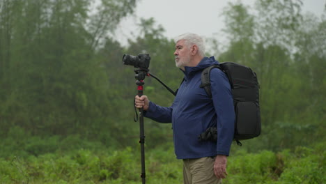 Alter-Mann-Geht,-Während-Er-Eine-Kamera-Mit-Einbeinstativ-Hält,-Im-Hintergrund-Der-Natur