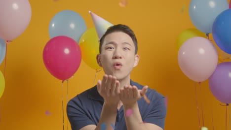 생일을 축하하는 파티 모자를 입은 남자의 스튜디오 초상화 카메라에 종이 콘페티를 불어넣는