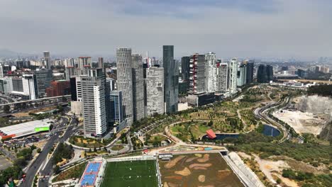 Flying-in-front-of-tall-buildings-at-the-La-Mexicana-park-in-sunny-Santa-Fe,-Mexico---Aerial-view