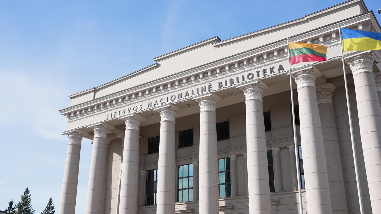Grandangolo della Biblioteca Nazionale Lituana Martynas Mažvydas, facciata neoclassica e colonne imponenti sotto un cielo azzurro e limpido, bandiere lituana e ucraina che sventolano dolcemente alla luce del giorno.