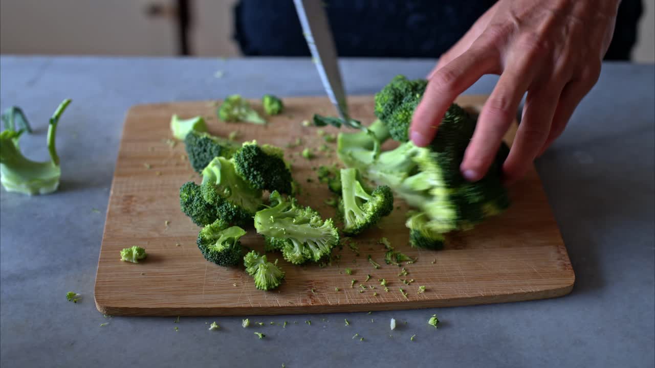 Primo piano di mani che tagliano fiori di broccoli verdi freschi su un tagliere di legno con un coltello affilato, con pezzi sparsi