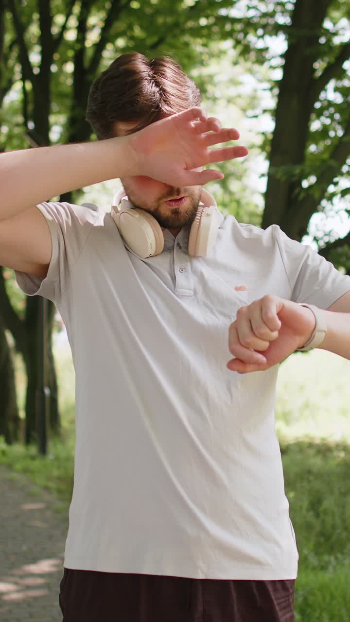 Giovane uomo attivo che fa jogging in un parco pubblico, guarda lo smartwatch e si riposa perché è stanco