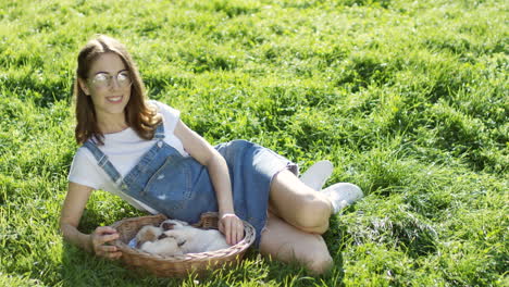 caucasian woman in glasses lying on the grass and petting labrador puppies sleeping in a basket in the park