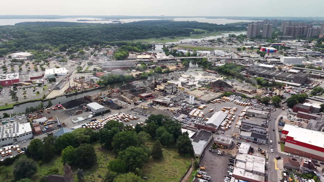 Expansive aerial view showcasing an industrial district with a prominent recycling plant and scrapyard. Contrasting with the lush green cemetery and the surrounding urban cityscape of the bronx