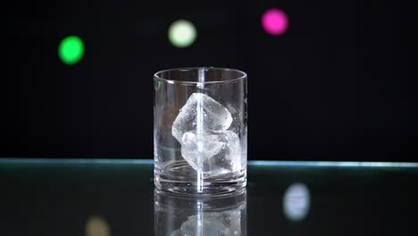 cubos de hielo gigantes cayendo en un vaso de licor sentado encima de una superficie de vidrio con un fondo negro con luces de hadas