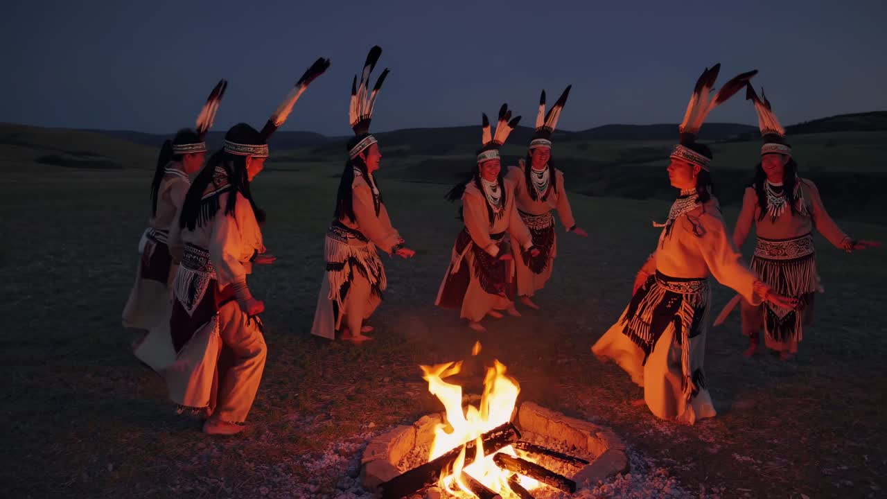 Eine ruhige Videoszene von Menschen in traditioneller Kleidung um ein Lagerfeuer in der Dämmerung, aus niedriger Perspektive aufgenommen.