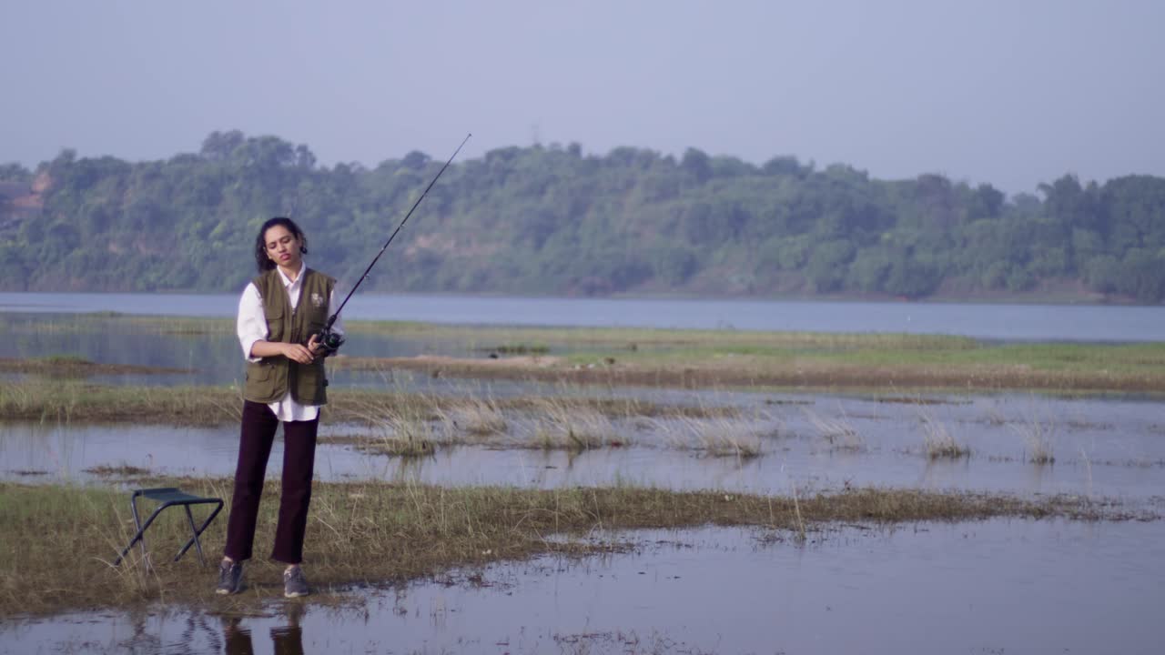 Seorang gadis Asia Selatan mengenakan rompi luar ruangan, berdiri dan melempar pancing ke air.