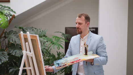 artist with color palette works in studio. thoughtful man in blue jacket draws applying pigment smears onto canvas on easel. professional painter job