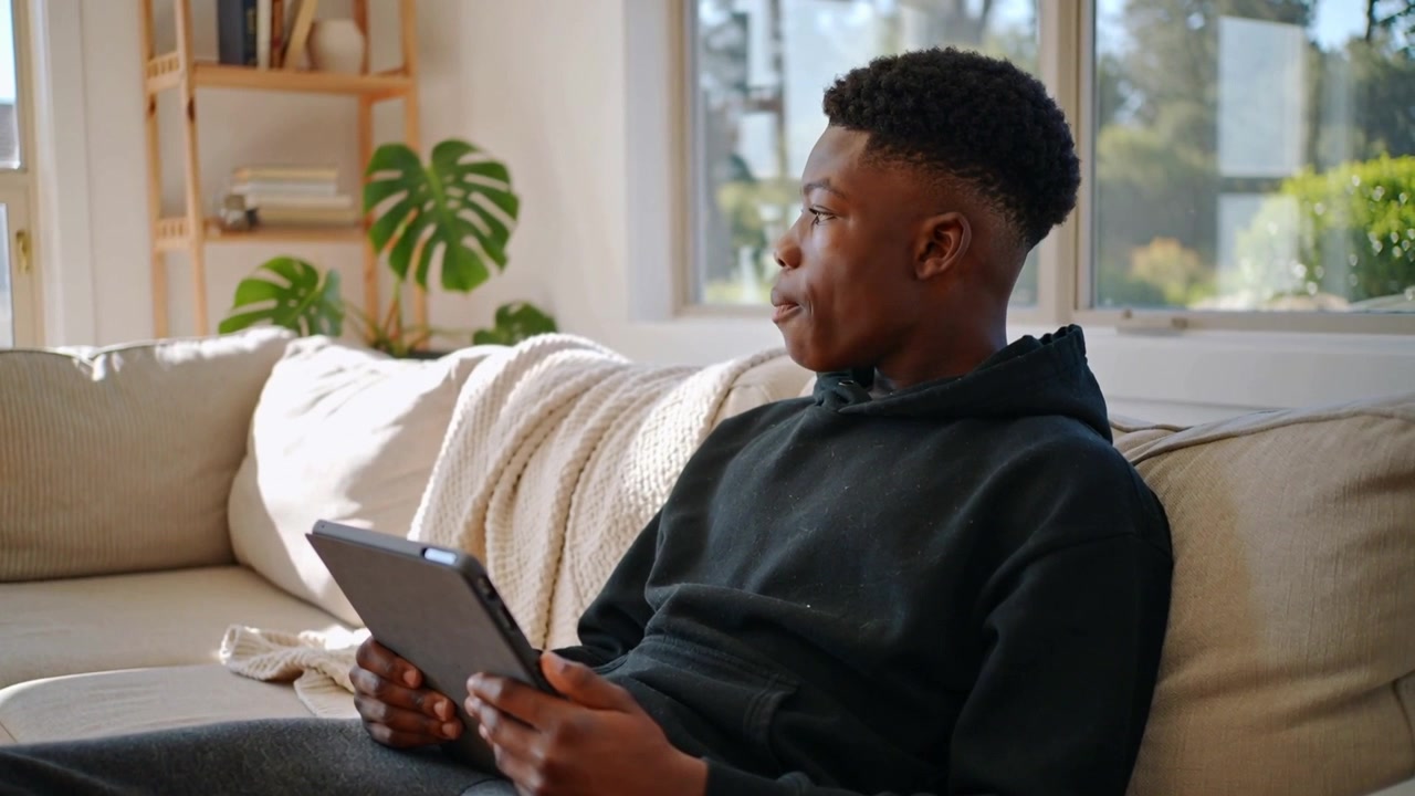 Noticing off-screen sound teen in black hoodie turning head on sofa by sunny window, holding tablet