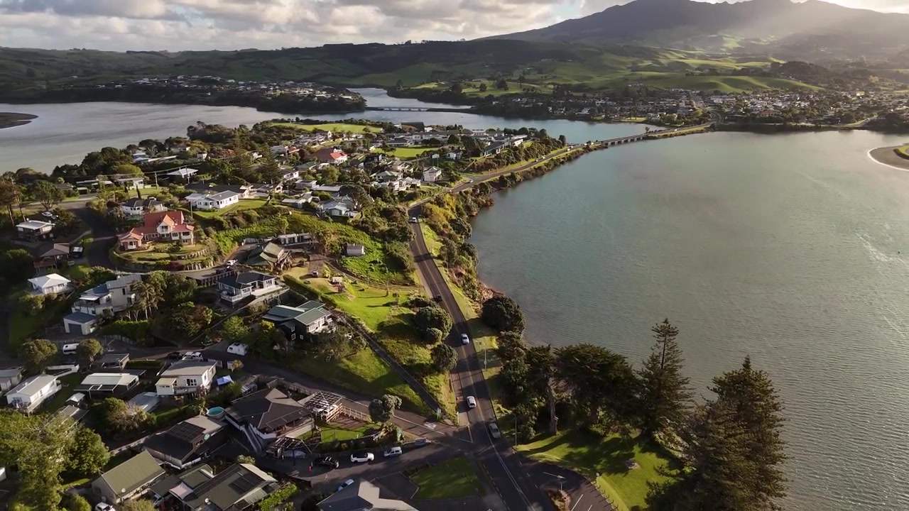 Splendide riprese aeree in 4K a 60fps che volano all'indietro sopra la città di Raglan, sulla costa occidentale della Nuova Zelanda. Questa fluida ripresa con drone all'indietro cattura il bellissimo insediamento costiero immerso nella calda luce del tramonto.